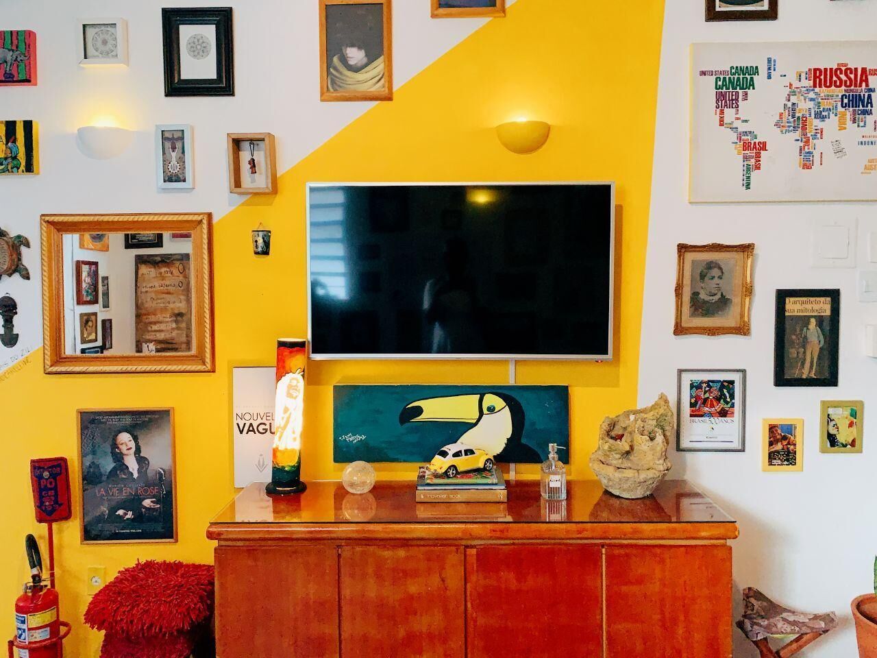 A room with a large flat-screen TV mounted on a yellow wall, surrounded by various framed pictures and artworks.