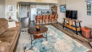 A living room with a sofa, a coffee table, a TV, and a kitchen in the background.