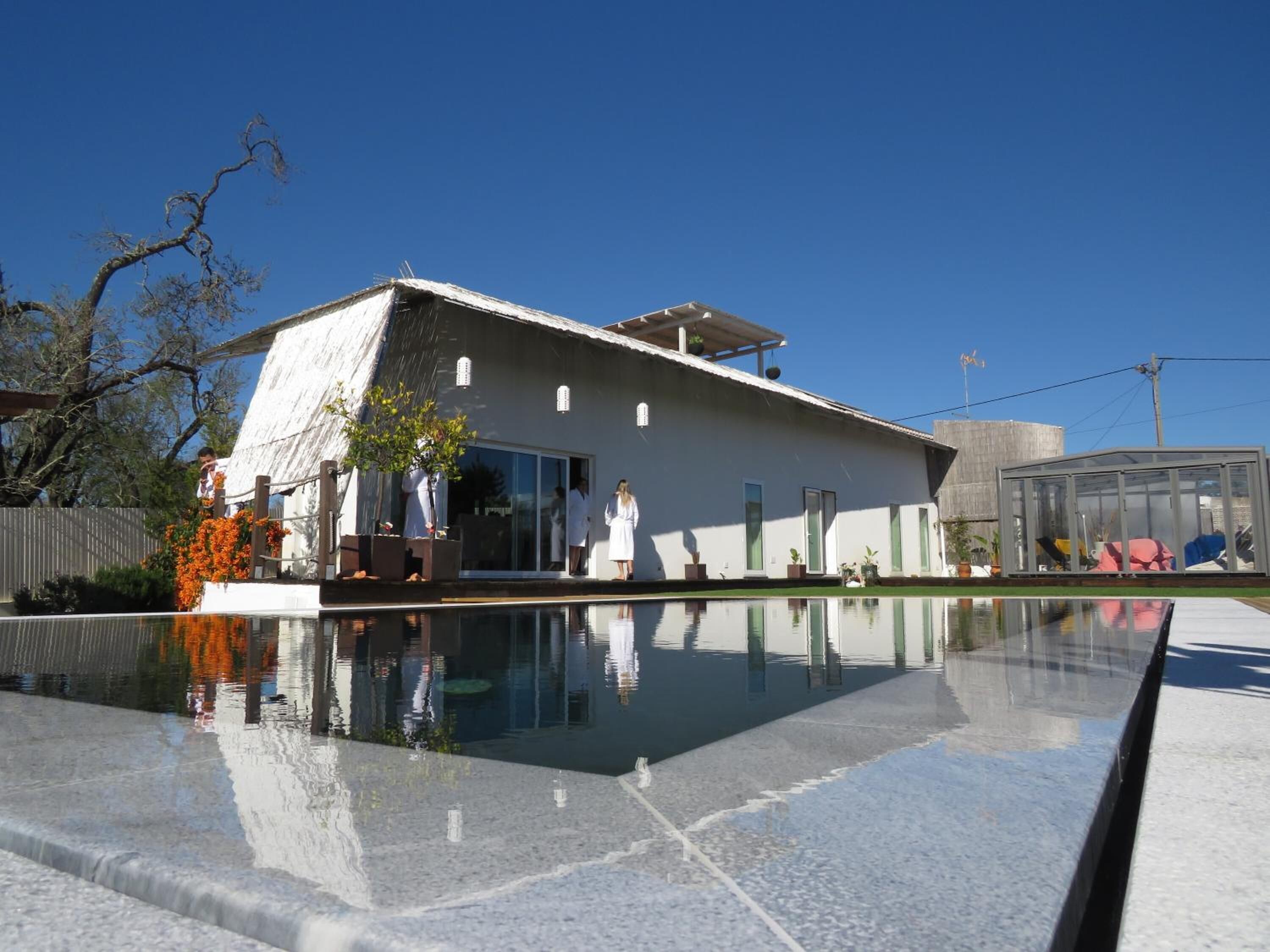 Un edificio moderno con un amplio ventanal, una piscina en primer plano y un cielo azul despejado.