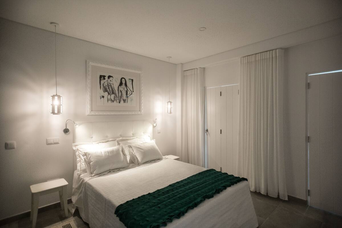 A bedroom with a bed, bedside lamps, a framed artwork, a green throw blanket, and a door with curtains.