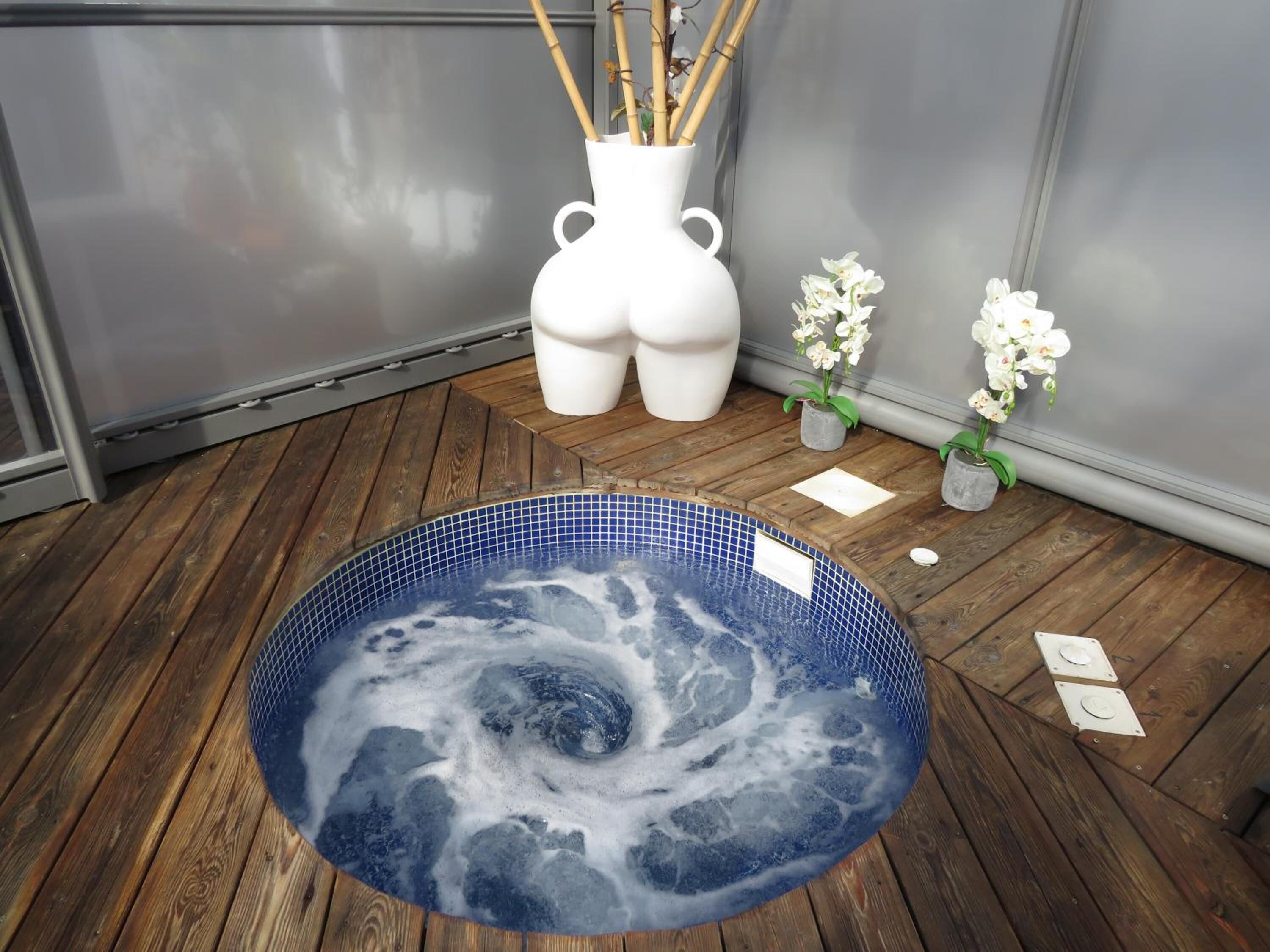 Ein Whirlpool mit blauem Mosaikfuß, umgeben von Holzboden, weißen Vasen und weißen Orchideen.