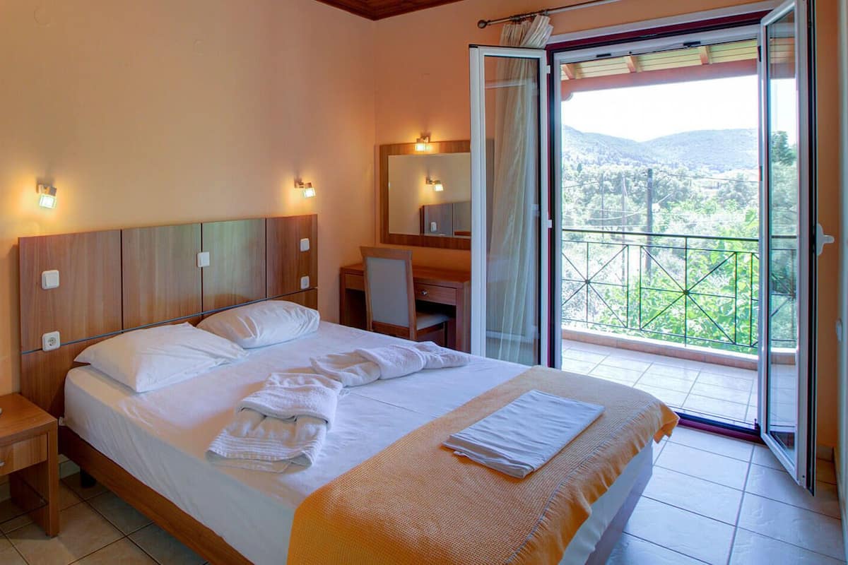 A bedroom with a bed, bedside table, and a desk with a chair. There is a balcony with a view of greenery and hills.