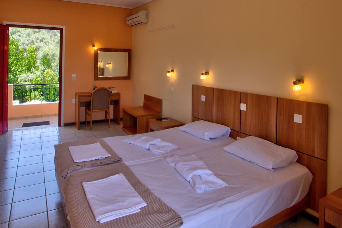 A hotel room with a large bed, a desk, a chair, and a balcony with a view of trees.