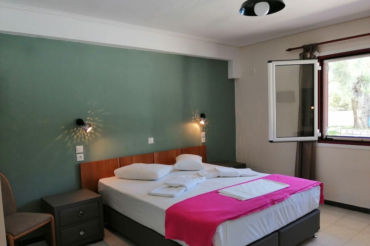 A neatly made bed with white linens and a pink blanket, a bedside table with a lamp, and a window with a view of greenery.