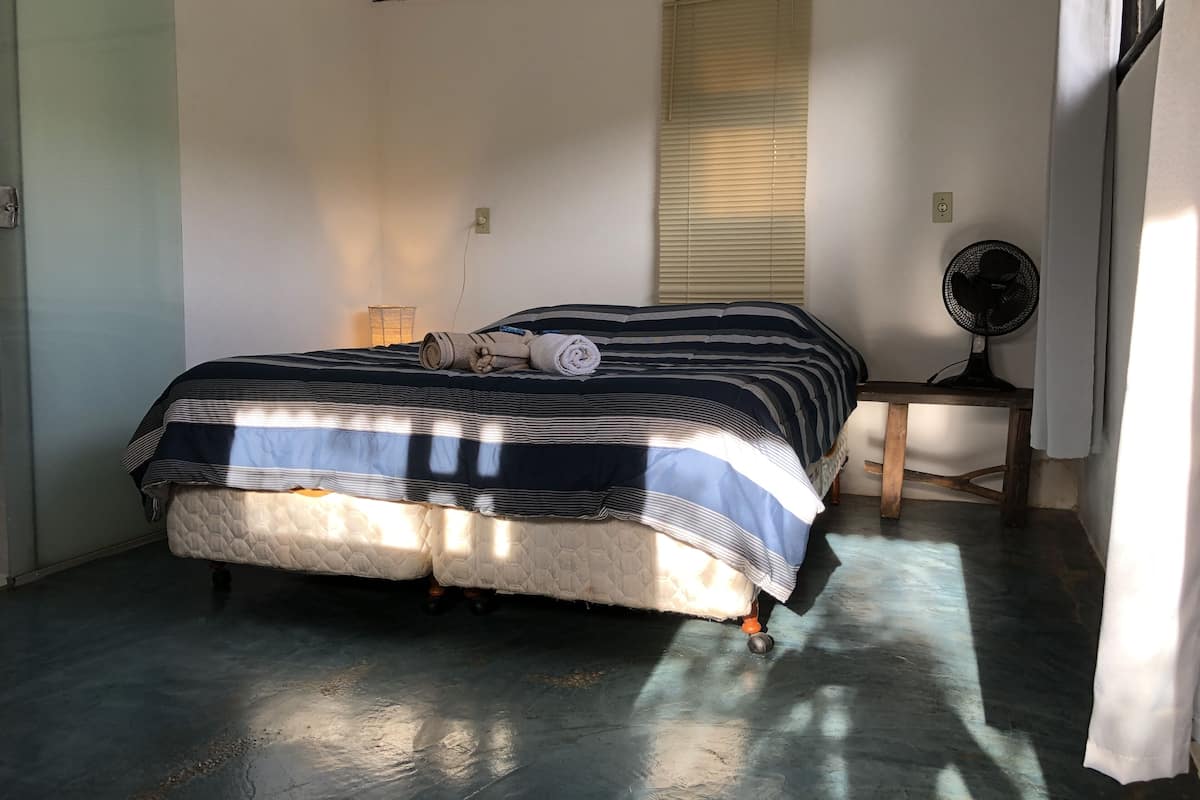 A bedroom with a bed, a bedside table with a fan, a window with blinds, and a lamp.