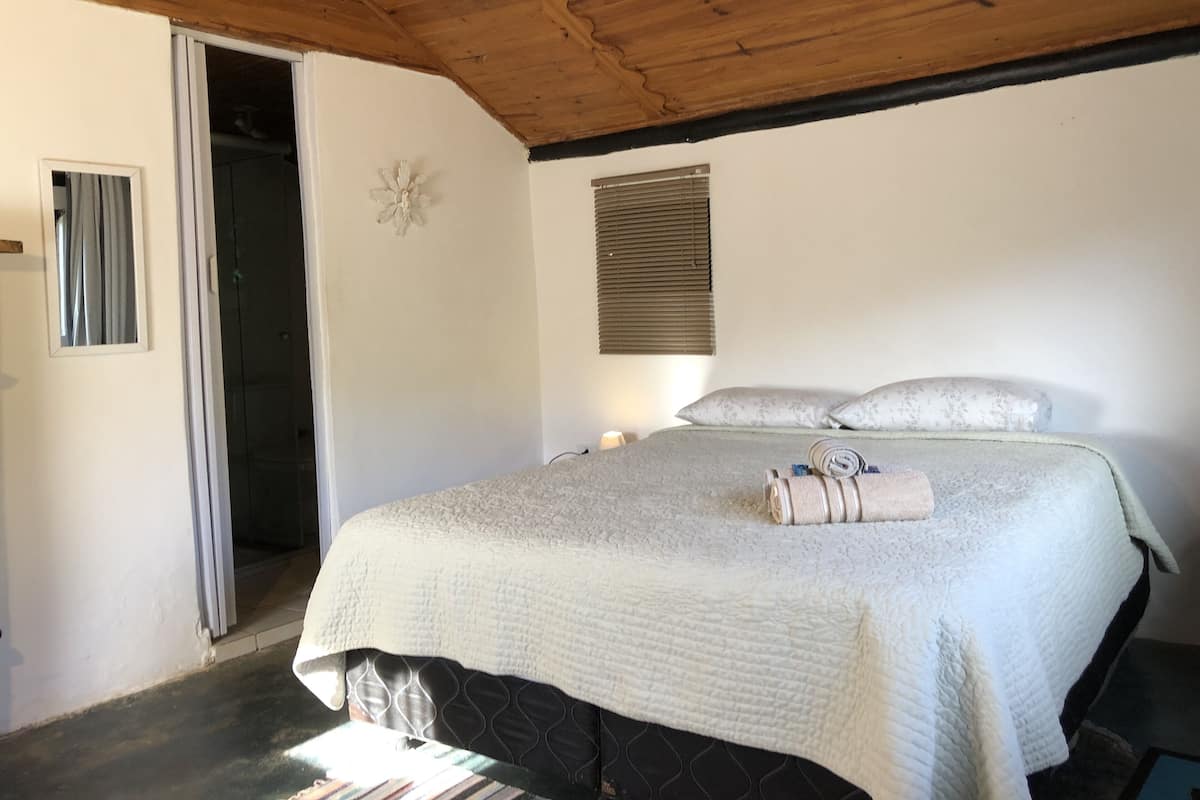 A bedroom with a wooden ceiling, a bed with a quilted cover, a bedside lamp, and a window with blinds.
