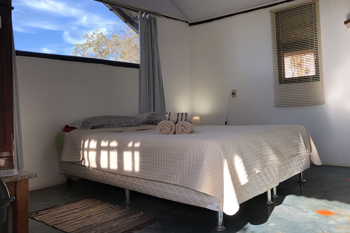 A bedroom with a bed, a window with blinds, a bedside lamp, and a power outlet.