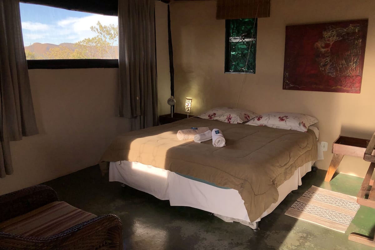 A bedroom with a bed, a chair, a window with curtains, a bedside lamp, and a painting on the wall.