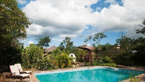 Una piscina con lettini, circondata da vegetazione lussureggiante e una struttura con tetto di paglia.