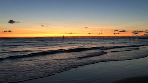 Una playa con olas al atardecer, un muelle que se adentra en el agua y una persona de pie sobre el muelle.