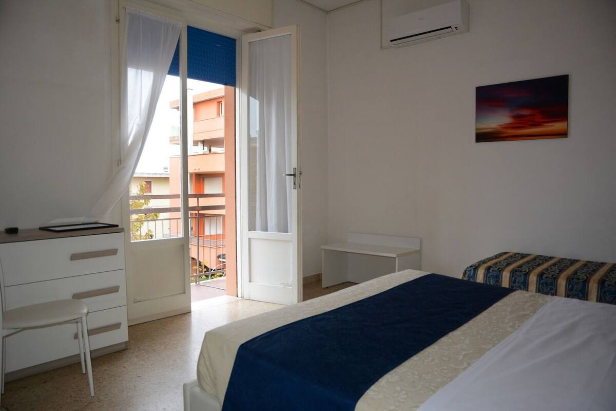 Ein Schlafzimmer mit Bett, Schreibtisch und Stuhl. Durch die Tür ist ein Balkon zu sehen.