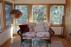 A cozy room with a wicker sofa, a glass coffee table, and a large window with a view of trees.