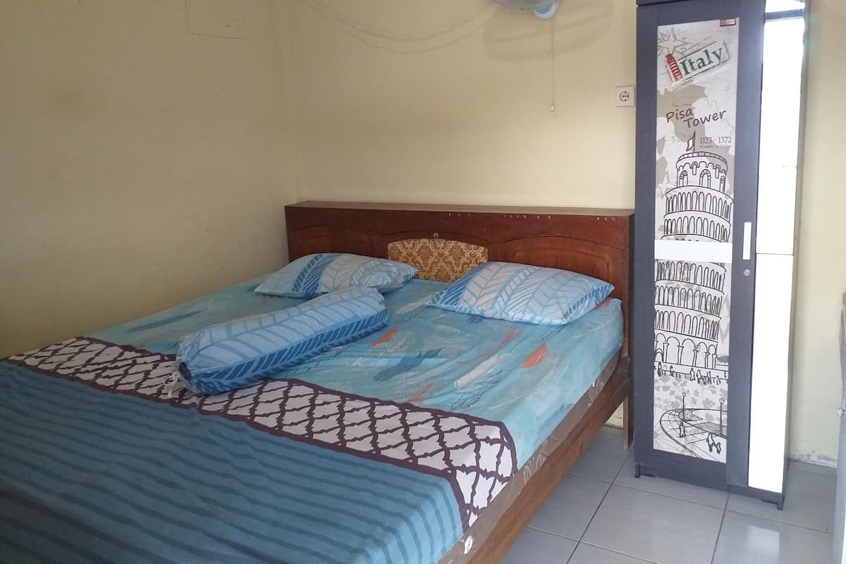 Une chambre à coucher avec un lit, une tête de lit en bois, un ventilateur mural et une porte ornée d’une affiche représentant la tour de Pise, en Italie.
