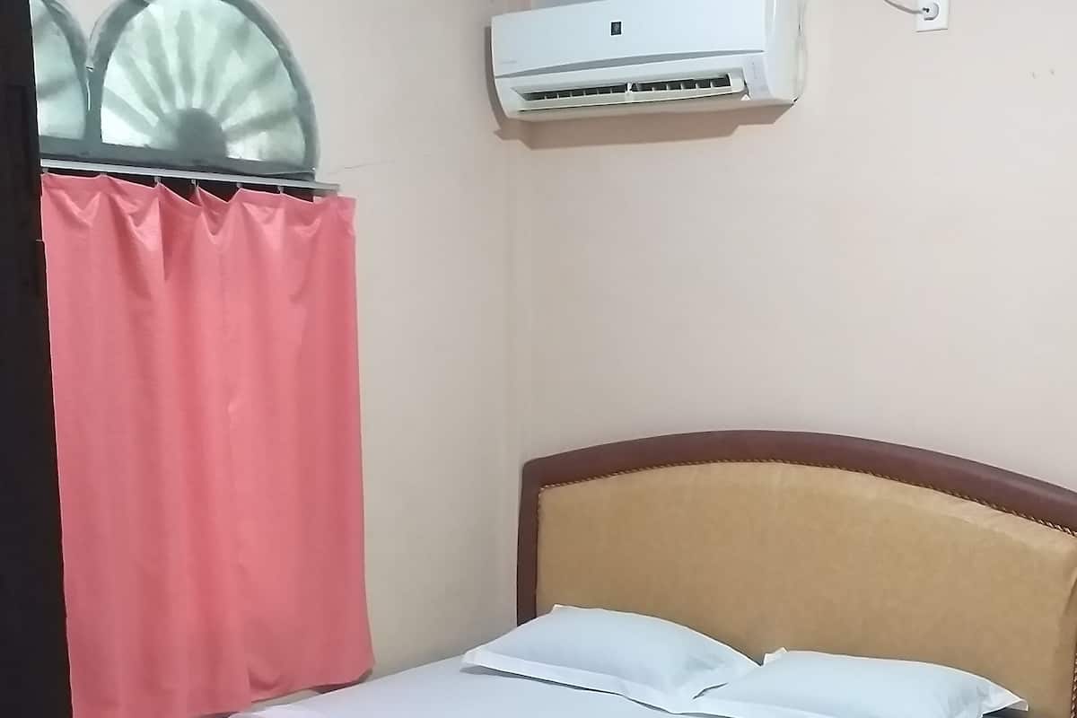 Un lit bien fait, avec du linge de lit blanc et une tête de lit marron, un climatiseur fixé au mur et un rideau rose, dont on aperçoit une partie.