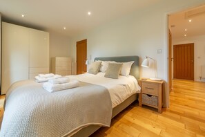 A neatly made bed with a white comforter and pillows, a bedside table with a lamp, and a wooden floor. - This spacious, detached holiday home is just a short walk from the beach. (12 Brecon Close)