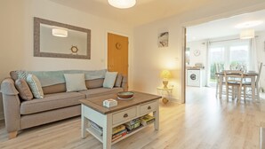 A living room with a sofa, coffee table, and bookshelves. There is a dining area with a table and chairs visible through an open door. - This property is an ideal, and very green choice for your family holiday. (32, Ashton Close)