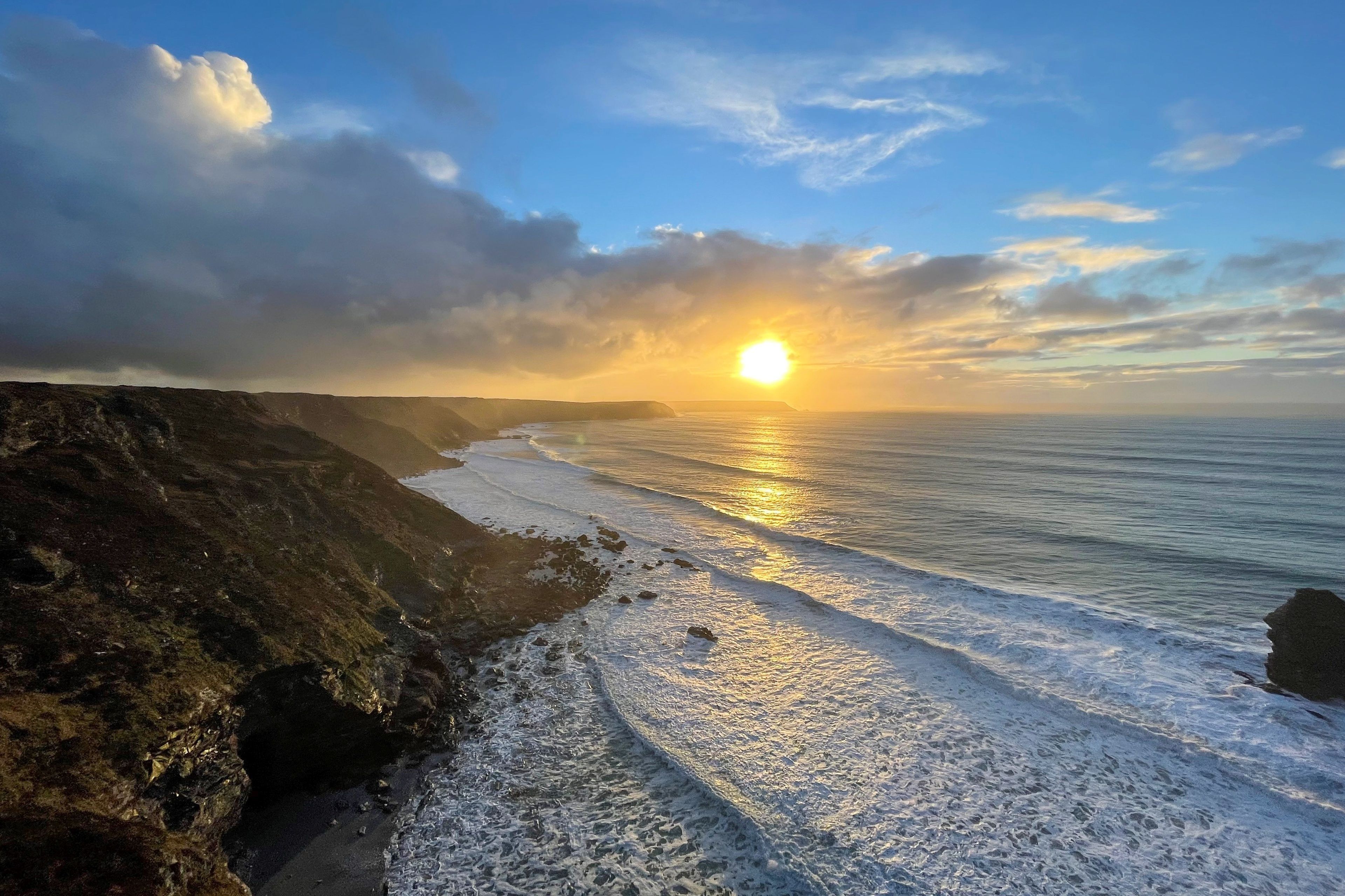 A coastal landscape with a sunset over the ocean, rocky cliffs, and a sandy beach.
