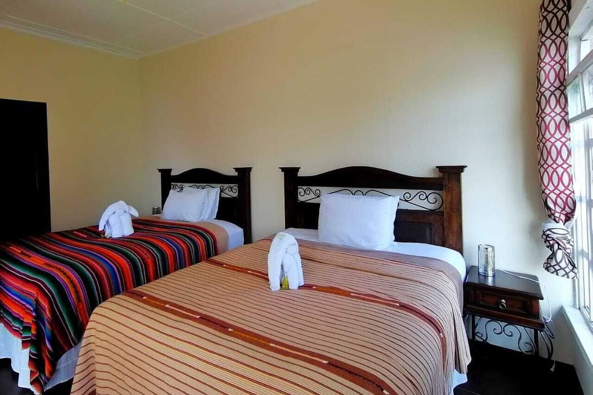 A hotel room with two beds, a ceiling fan, a window with patterned curtains, and a bedside table with a lamp.