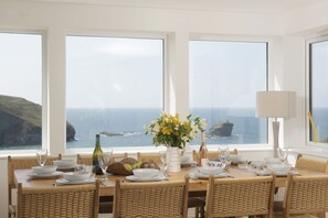 Mesa de jantar posta para seis com vista para o mar, cadeiras de vime e um vaso de flores.