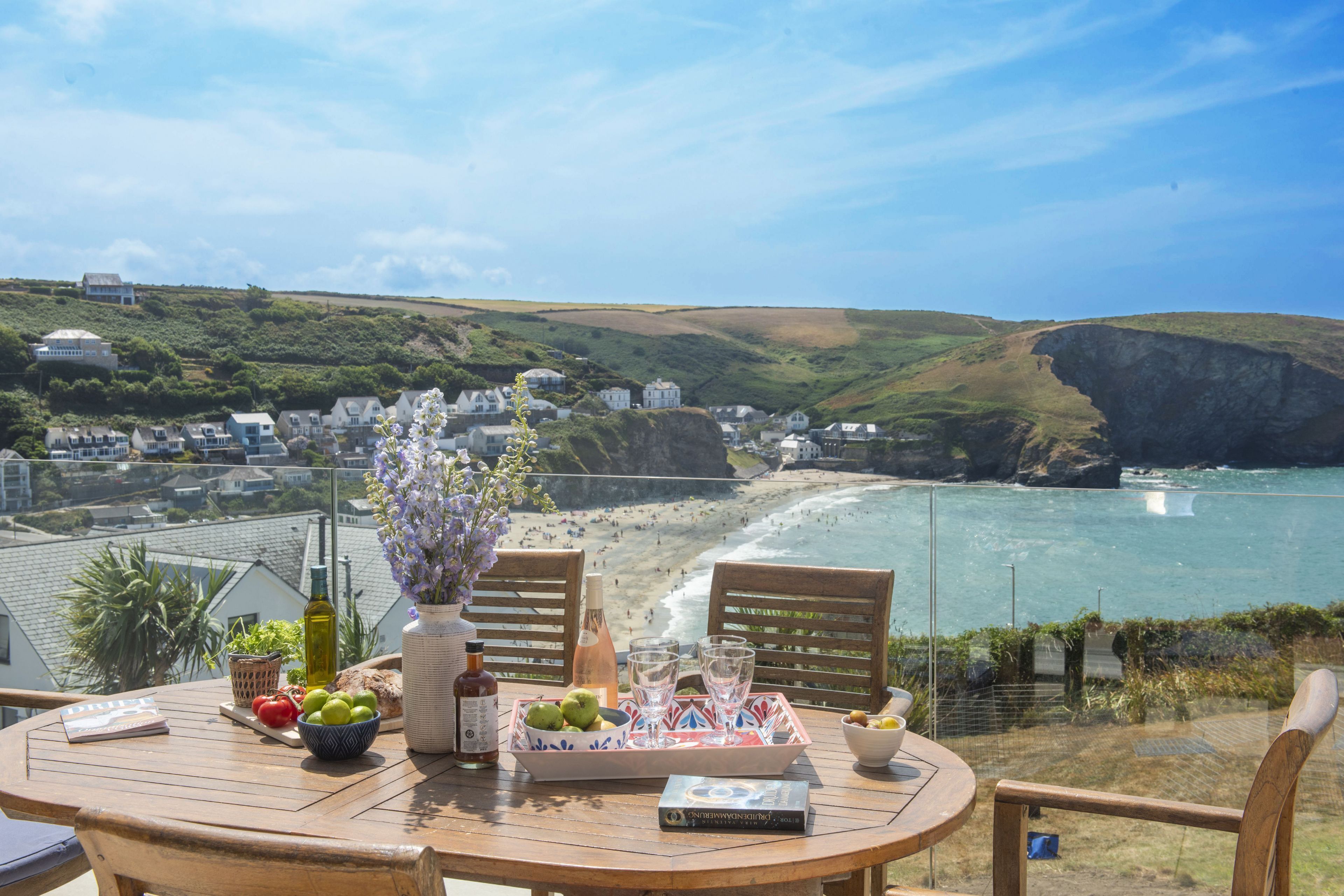 A wooden table with a bottle, glasses, and a bowl of fruit, overlooking a coastal town with houses and cliffs.