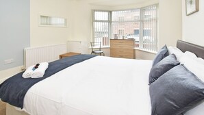 A neatly made bed with a dark blue blanket and pillows, a wooden dresser, a chair, and a window with a view of the outside. - Townhouse @ Earle Street Crewe (Crewe)