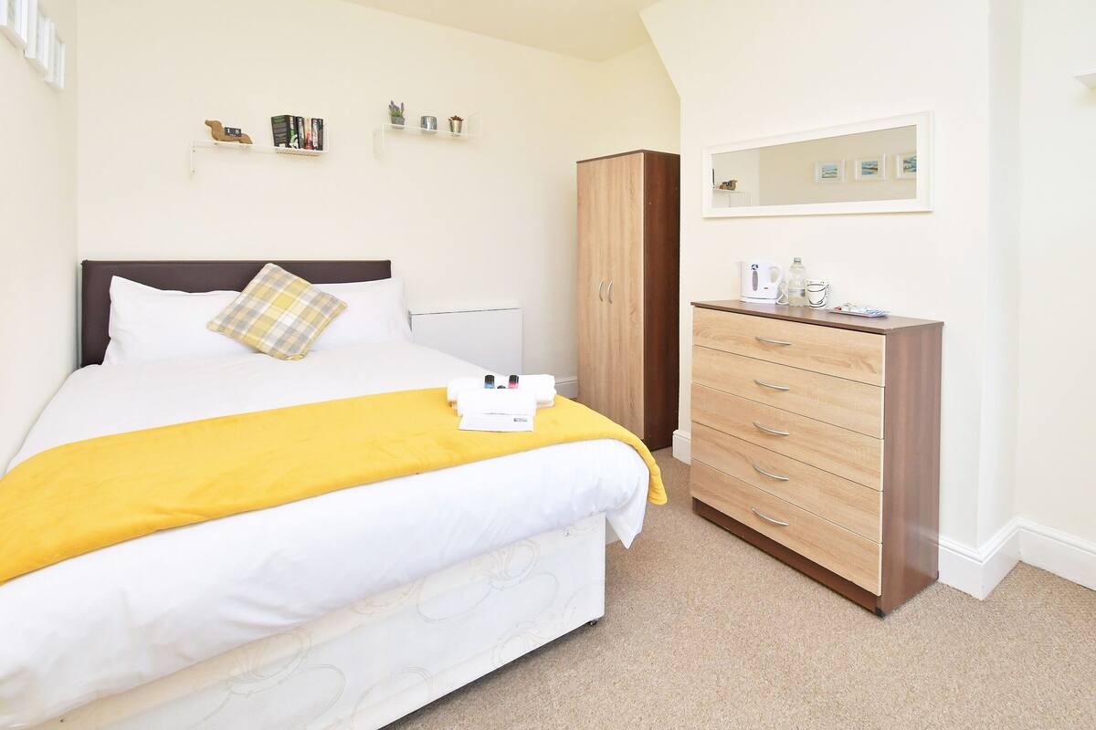 Un dormitorio ordenado con cama, armario, mesita de noche con espejo y estante con libros y objetos decorativos.