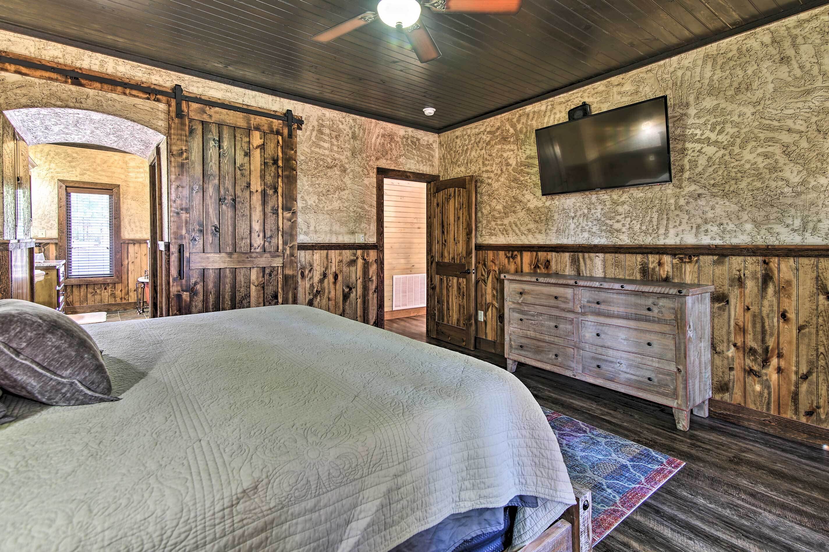 A bedroom with a large bed, wooden walls, a ceiling fan, a TV mounted on the wall, and a dresser.