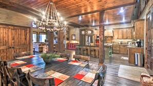 Un interior rústico de madera con una amplia mesa de comedor, sillas de madera, una araña y una zona de cocina.