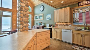 A kitchen with wooden cabinets, a stone accent wall, a clock, and a view of the water outside. - Hike & Ski! Family-Friendly Lake Mitchell Oasis (Cadillac)