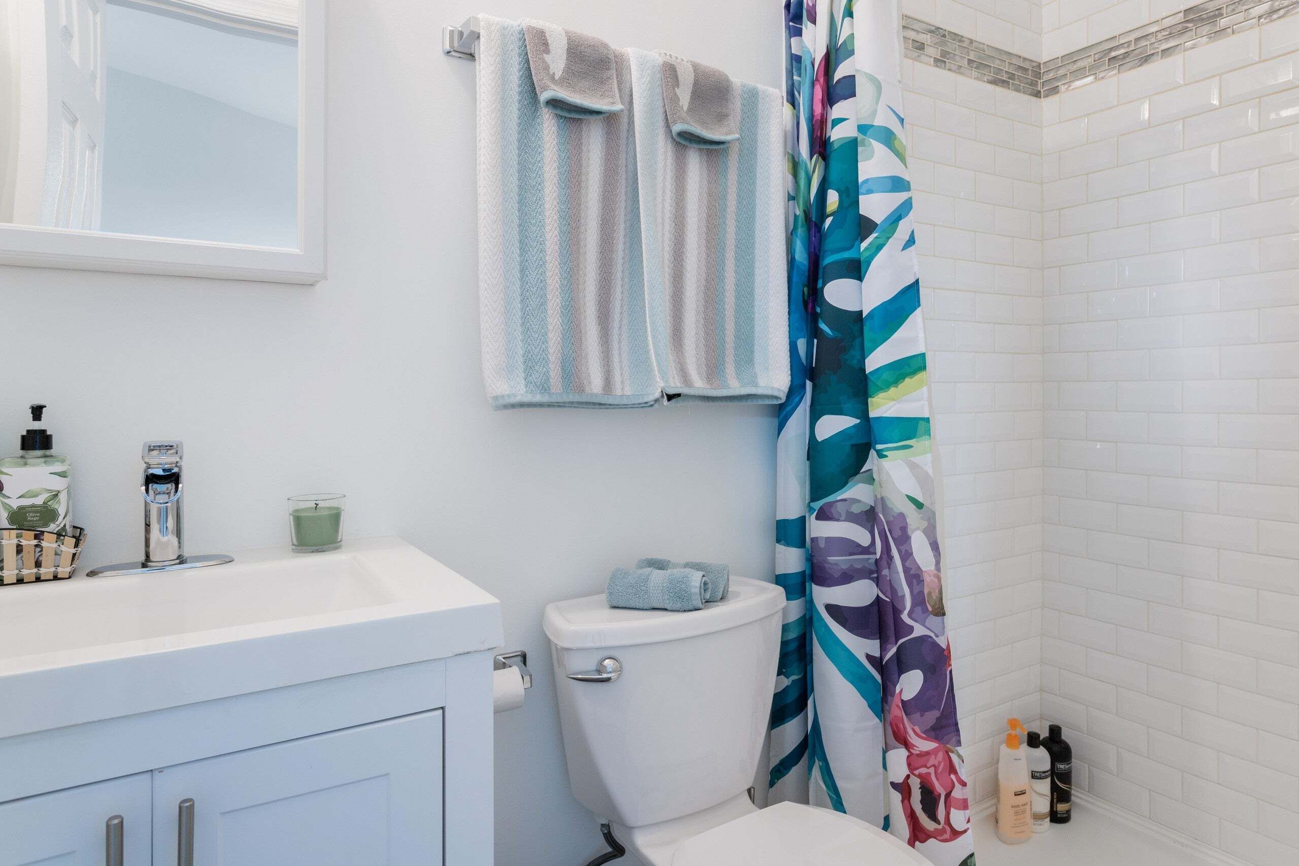 Une salle de bain avec un lavabo blanc, des toilettes et un rideau de douche orné d’un motif de feuilles. Deux serviettes sont accrochées au mur et une bouteille de shampoing est posée sur le sol.