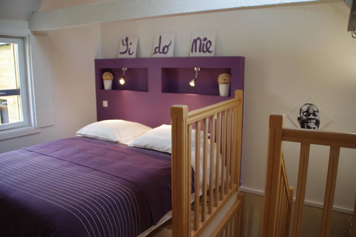 Un dormitorio con colcha morada, cabecera de madera y estanterías con adornos.