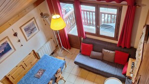 A cozy room with a wooden ceiling, a sofa with red cushions, a table with a blue tablecloth, and a window with red curtains.