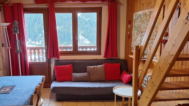 A cozy room with a wooden floor, a sofa with red and beige cushions, a small round table, and a window with red curtains.