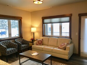 A living room with a sofa, two patterned armchairs, a coffee table, a lamp, and large windows with blinds. - ★Glacier Courtyards Condo ★ Walk Everywhere! (Columbia Falls)