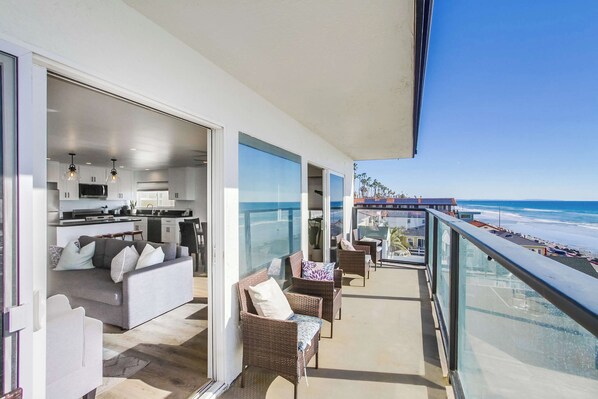 A balcony with a view of the beach, a living area with a sofa and chairs, and a kitchen area.