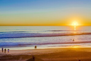 Escena de playa con el sol poniéndose sobre el océano, gente caminando por la orilla y un cielo despejado.