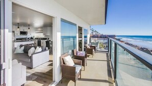 A balcony with a view of the beach, furnished with sofas and chairs, and a kitchen area visible through an open door. - The Pier House | A Luxury Oceanfront Penthouse (Oceanside)