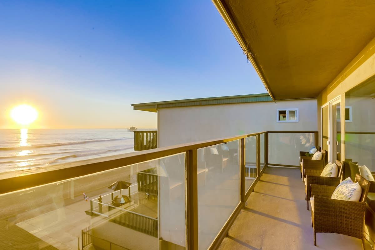 Varanda com vista para o oceano ao pôr do sol, mobiliário de vime e guarda-corpo de vidro.