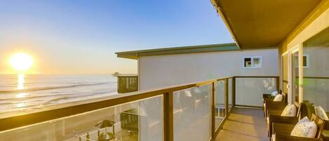 A balcony with a view of the ocean at sunset, furnished with wicker chairs and cushions.