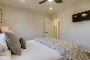 Une chambre à coucher avec un lit, des oreillers, un ventilateur de plafond, un téléviseur fixé au mur et une porte donnant sur une autre pièce.