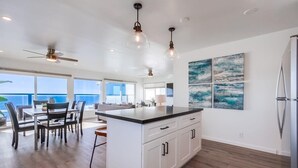 A spacious kitchen with a large island, white cabinetry, and pendant lighting. There are large windows offering a view of the ocean. - The Pier House | A Luxury Oceanfront Penthouse (Oceanside)
