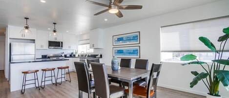 A modern kitchen with white cabinets, a central island, and bar stools. There is a dining area with a wooden table and chairs.