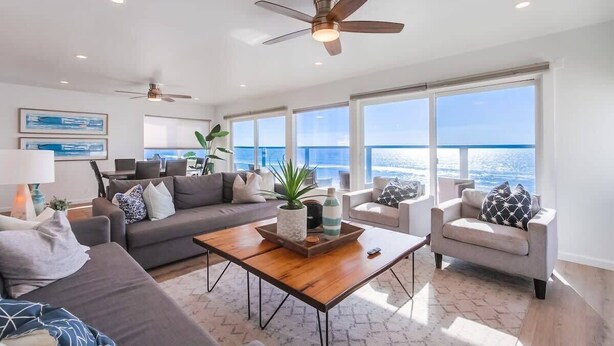 A modern living room with a large window, a ceiling fan, and a wooden coffee table. - The Pier House | A Luxury Oceanfront Penthouse (Oceanside)