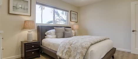 A bedroom with a bed, two bedside tables, a ceiling fan, and a window with a view of palm trees.