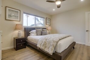 A bedroom with a bed, two bedside tables, a ceiling fan, and a window with a view of palm trees.