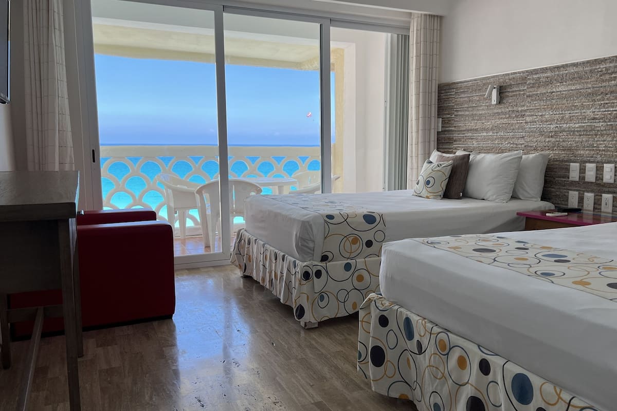 A hotel room with two beds, a red chair, a wooden floor, and a balcony with a view of the sea.