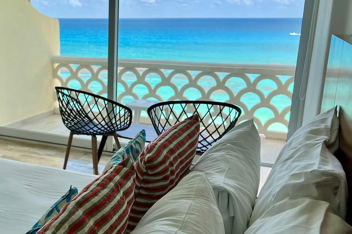 A bedroom with a view of the ocean, a bed with white linens, two black wireframe chairs, and a patterned pillow.