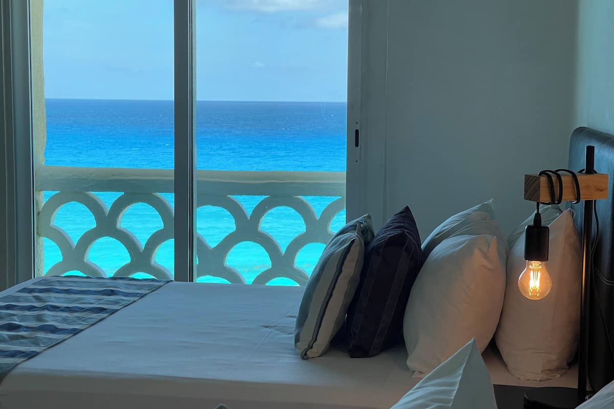 A bedroom with a bed, pillows, a bedside table with a lamp, and a view of the ocean through a balcony.