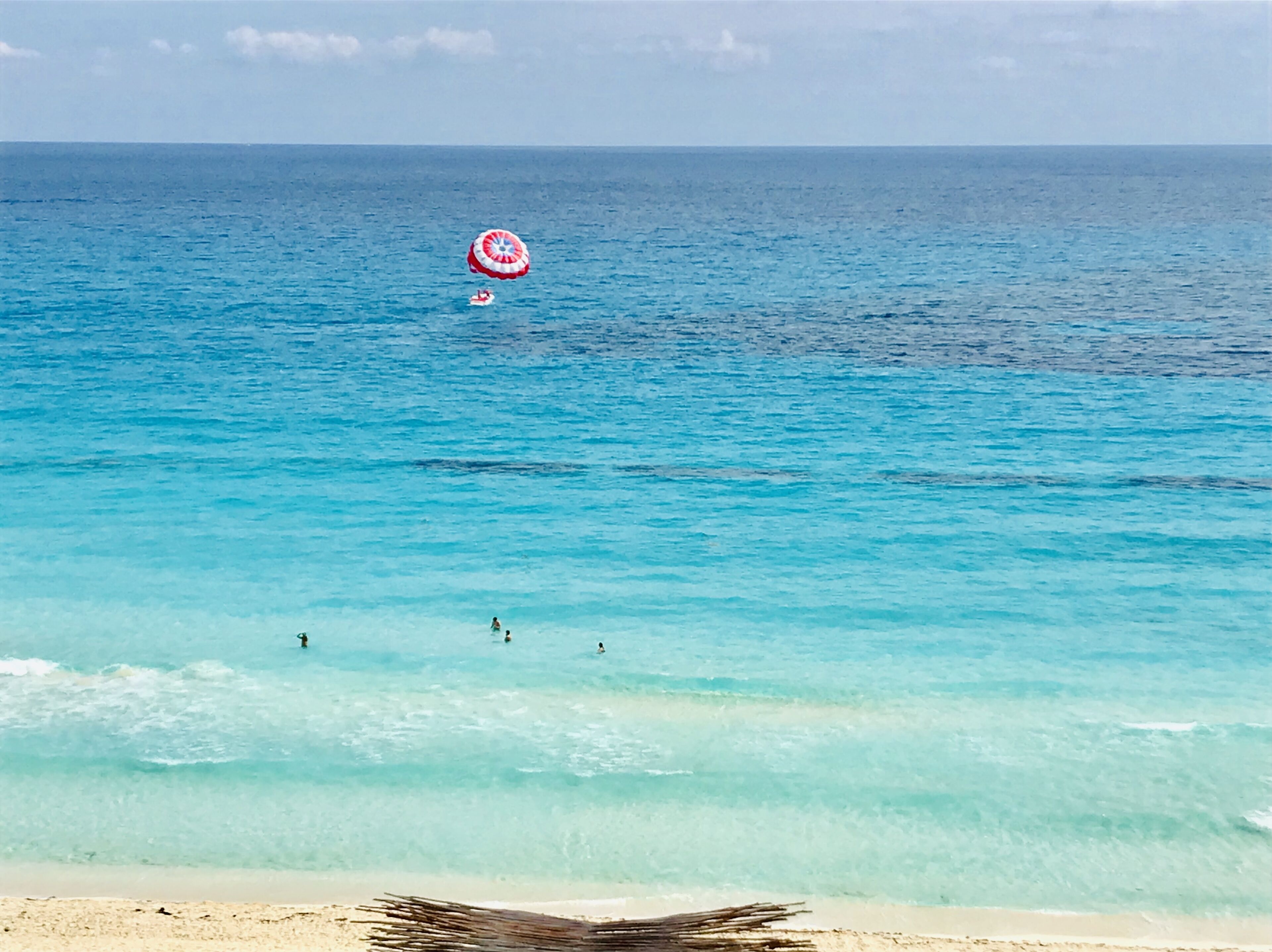 Uma praia com águas turquesa cristalinas, areal arenoso e um paraquedas a flutuar ao longe.