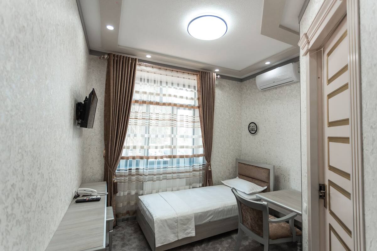 A compact hotel room with a single bed, a small desk with a chair, a flat-screen TV mounted on the wall, and a window with curtains.